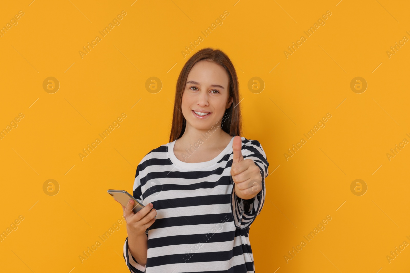 Photo of Portrait of teenage girl with smartphone showing thumb up on orange background