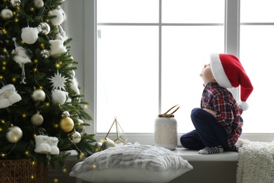 Cute little boy in Santa hat on window sill near Christmas tree at home