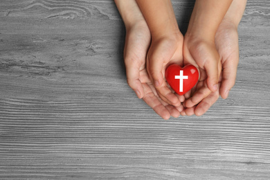 Woman and child holding heart with cross symbol on grey wooden background, top view with space for text. Christian religion