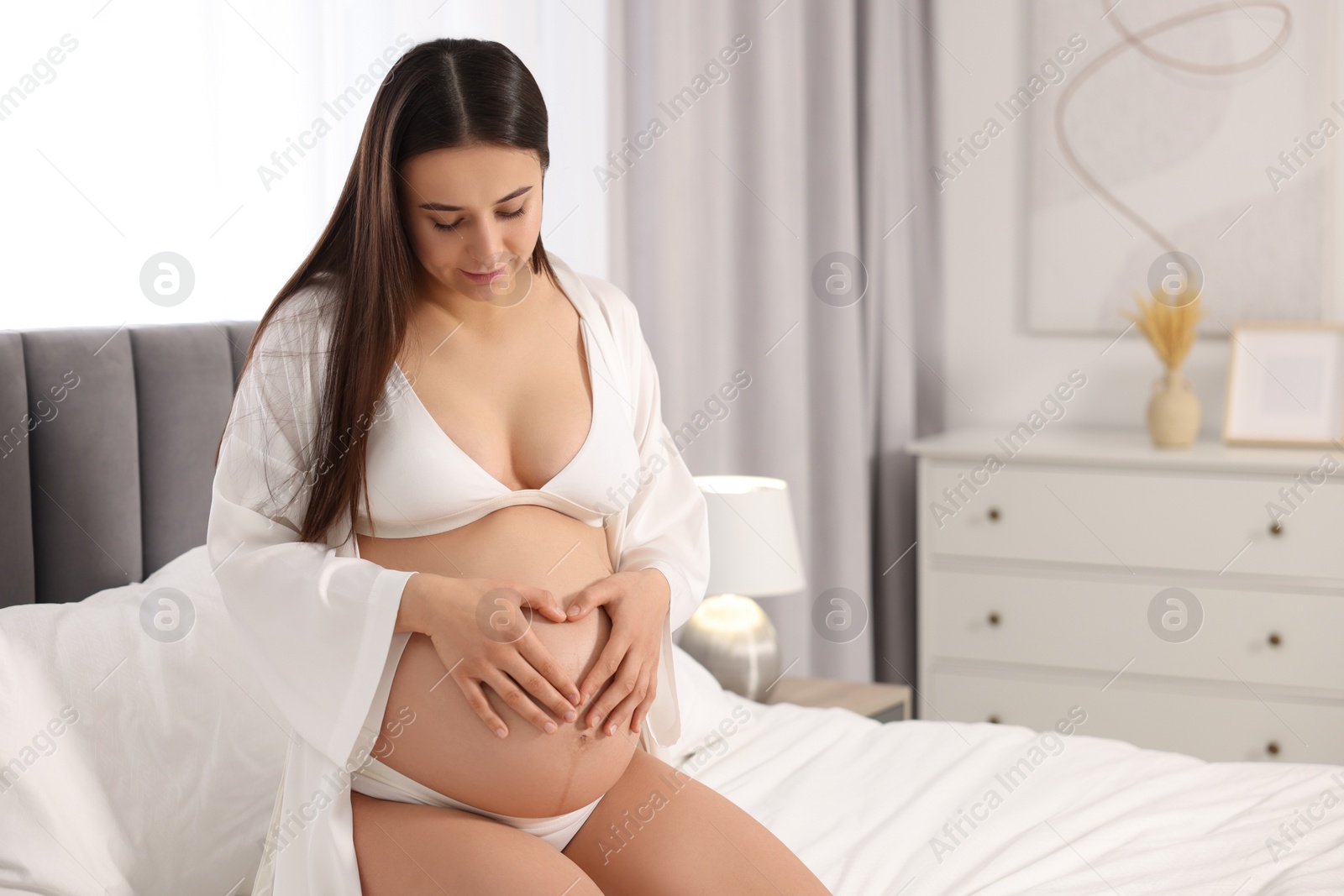 Photo of Beautiful pregnant woman in stylish comfortable underwear and robe on bed at home, space for text