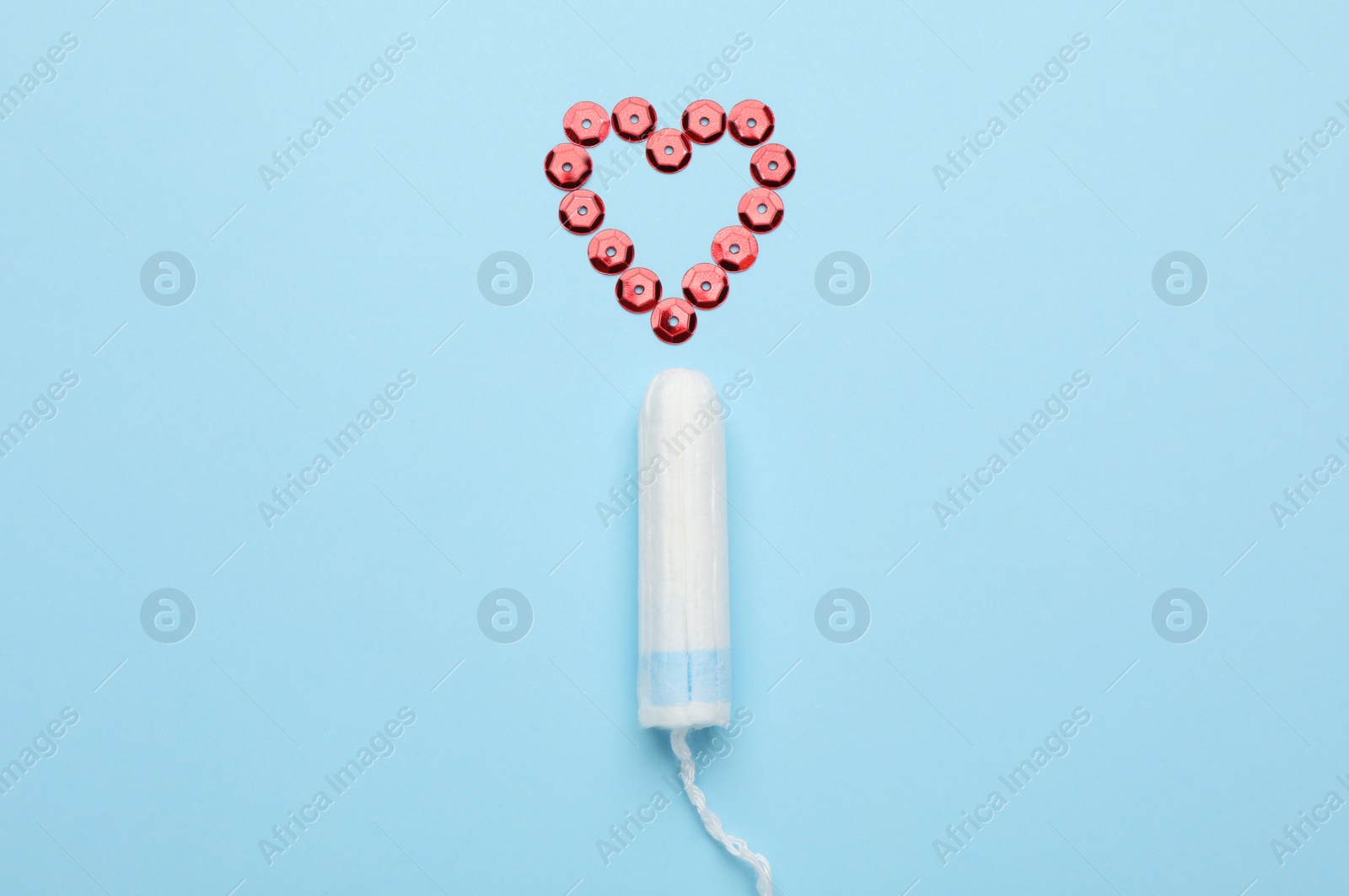 Photo of Tampon near heart made of red sequins on light blue background, flat lay. Menstrual hygiene product