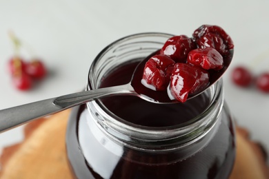 Photo of Spoon of delicious pickled cherries over jar, closeup