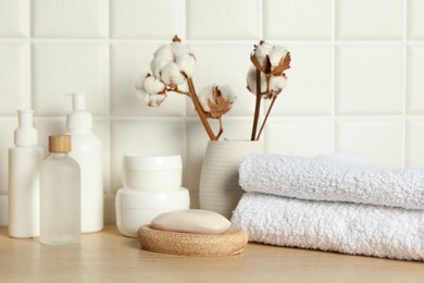 Photo of Different bath accessories, personal care products and cotton flowers in vase on wooden table near white tiled wall