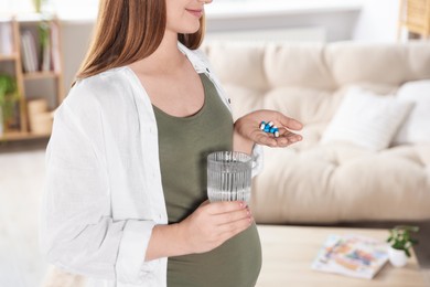 Photo of Pregnant woman holding pile of pills and glass with water at home, closeup