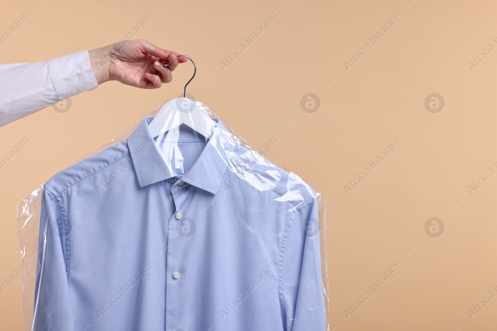 Photo of Dry-cleaning service. Woman holding shirt in plastic bag on beige background, closeup. Space for text