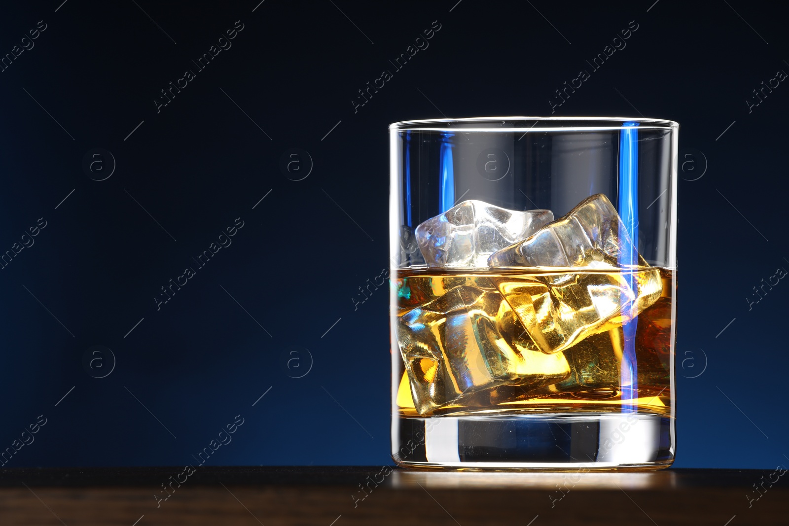 Photo of Whiskey with ice cubes in glass on table against blue background, closeup. Space for text