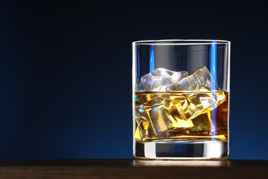 Photo of Whiskey with ice cubes in glass on table against blue background, closeup. Space for text