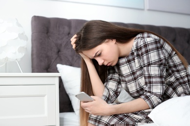Upset teenage girl with smartphone sitting on bed. Space for text