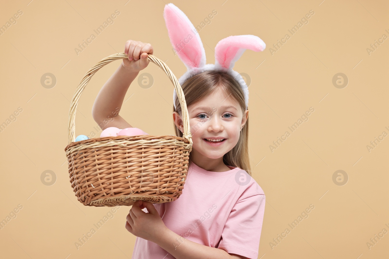 Photo of Easter celebration. Cute girl with bunny ears holding basket of painted eggs on beige background
