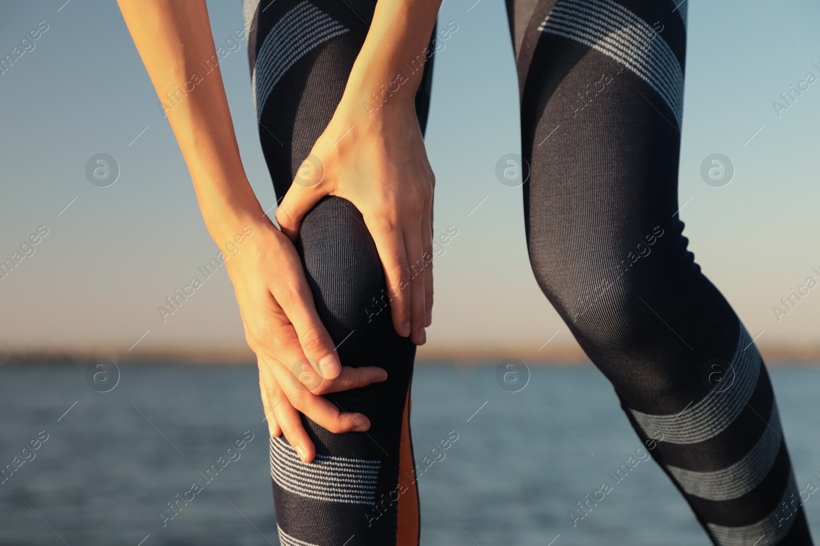 Photo of Young woman in sportswear having knee problems near river at sunset, closeup