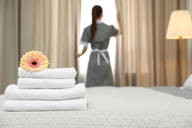 Photo of Stack of clean towels with flower on bed and blurred chambermaid in hotel room, closeup. Space for text