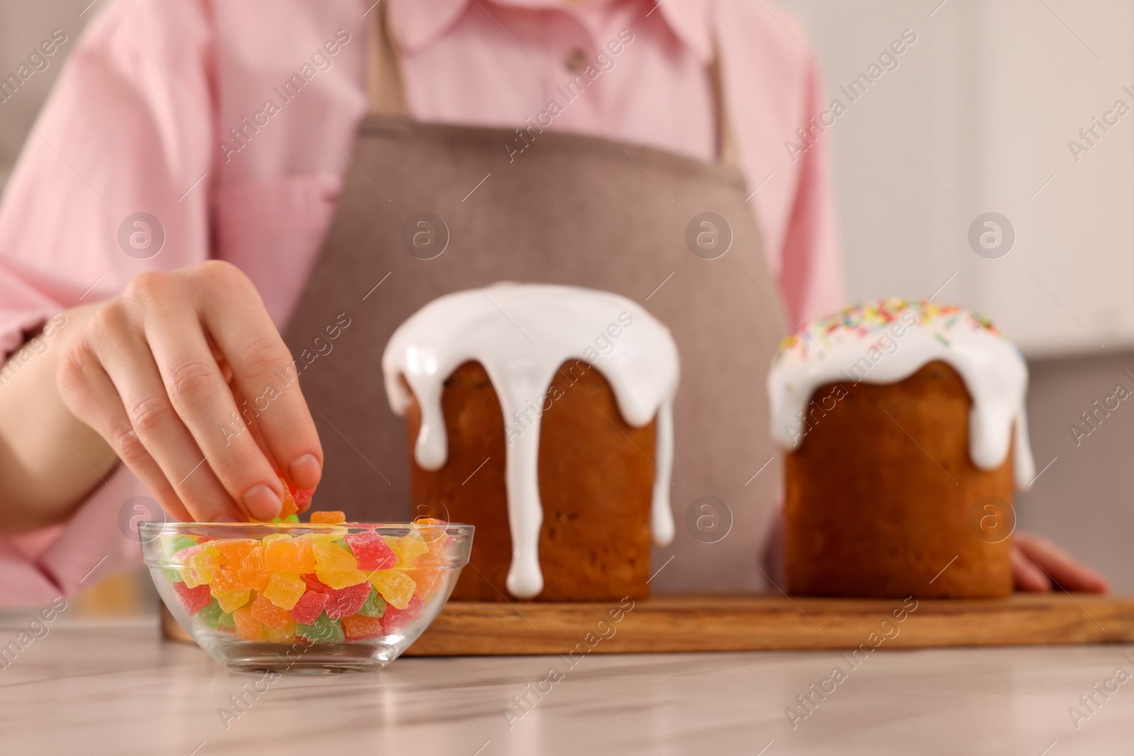 Photo of Woman decorating delicious Easter cake with pieces of candied fruits at white marble table in kitchen, closeup