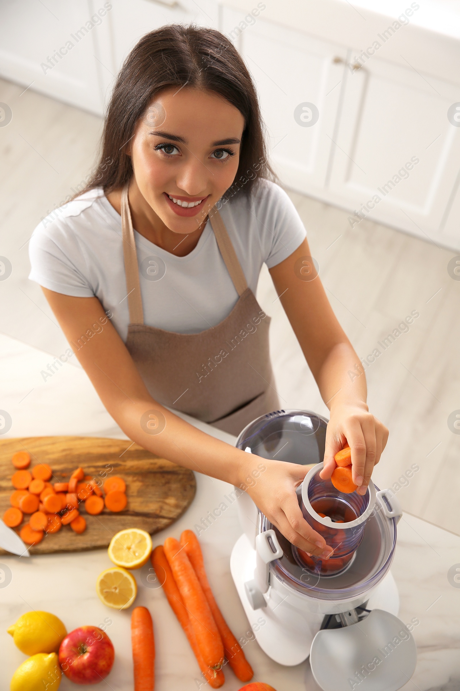 Photo of Young woman putting fresh slices of carrot into juicer at table in kitchen, above view