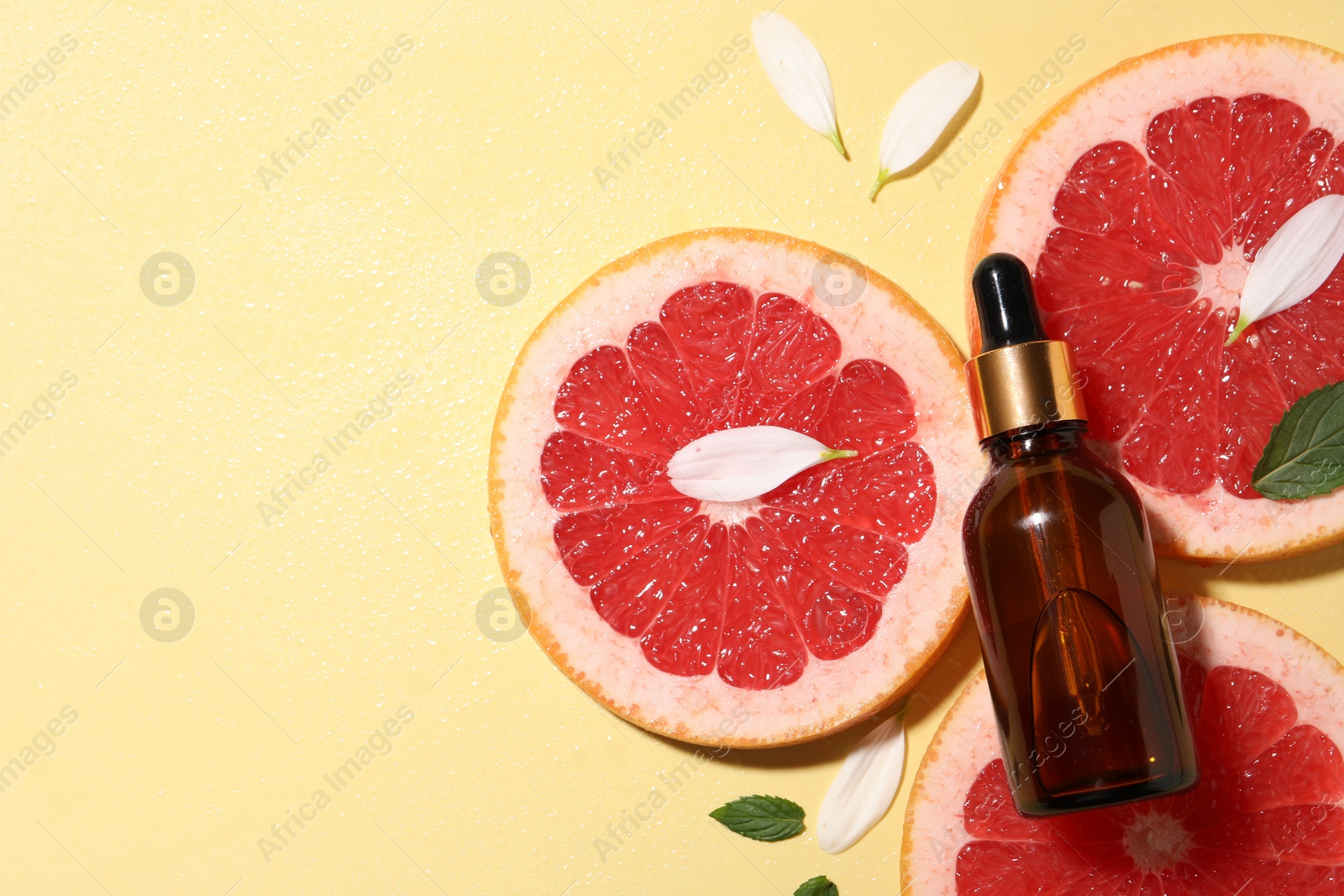 Photo of Bottle of cosmetic serum, grapefruit slices and green leaves on wet yellow background, flat lay. Space for text