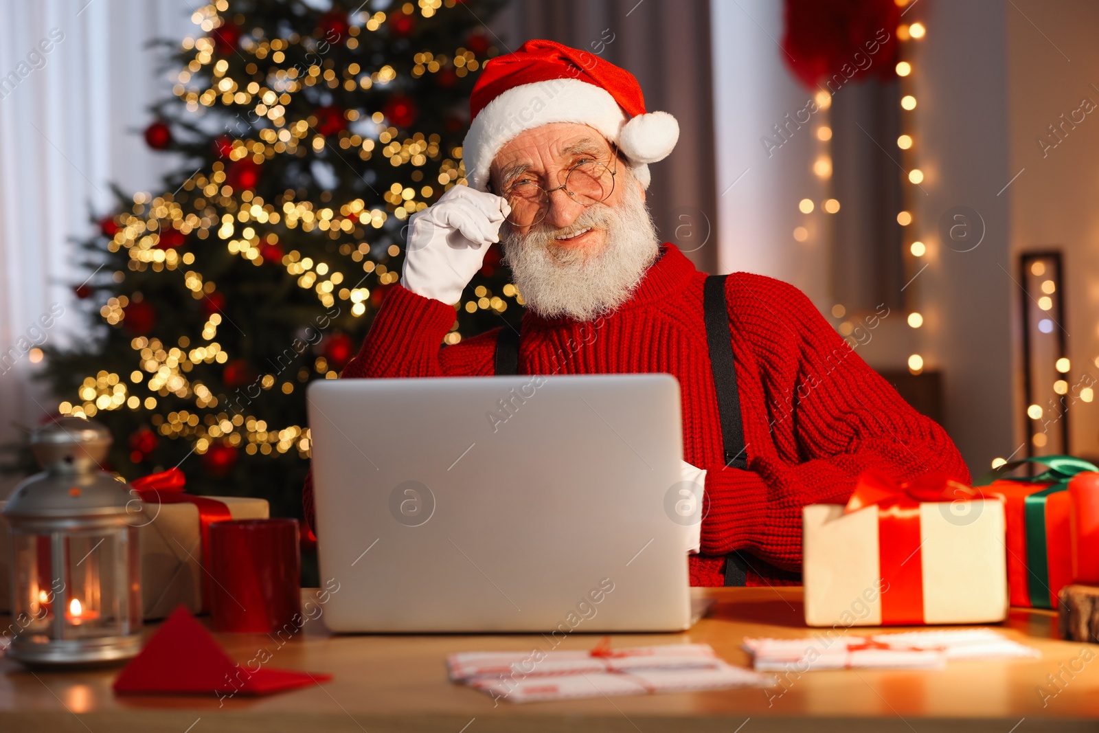 Photo of Santa Claus using laptop and laughing at his workplace in room with Christmas tree