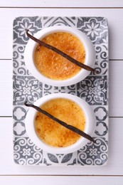 Delicious creme brulee in bowls and vanilla pods on white wooden table, top view
