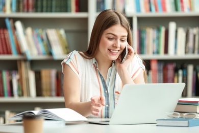 Photo of Young woman talking on phone and working with laptop at table in library