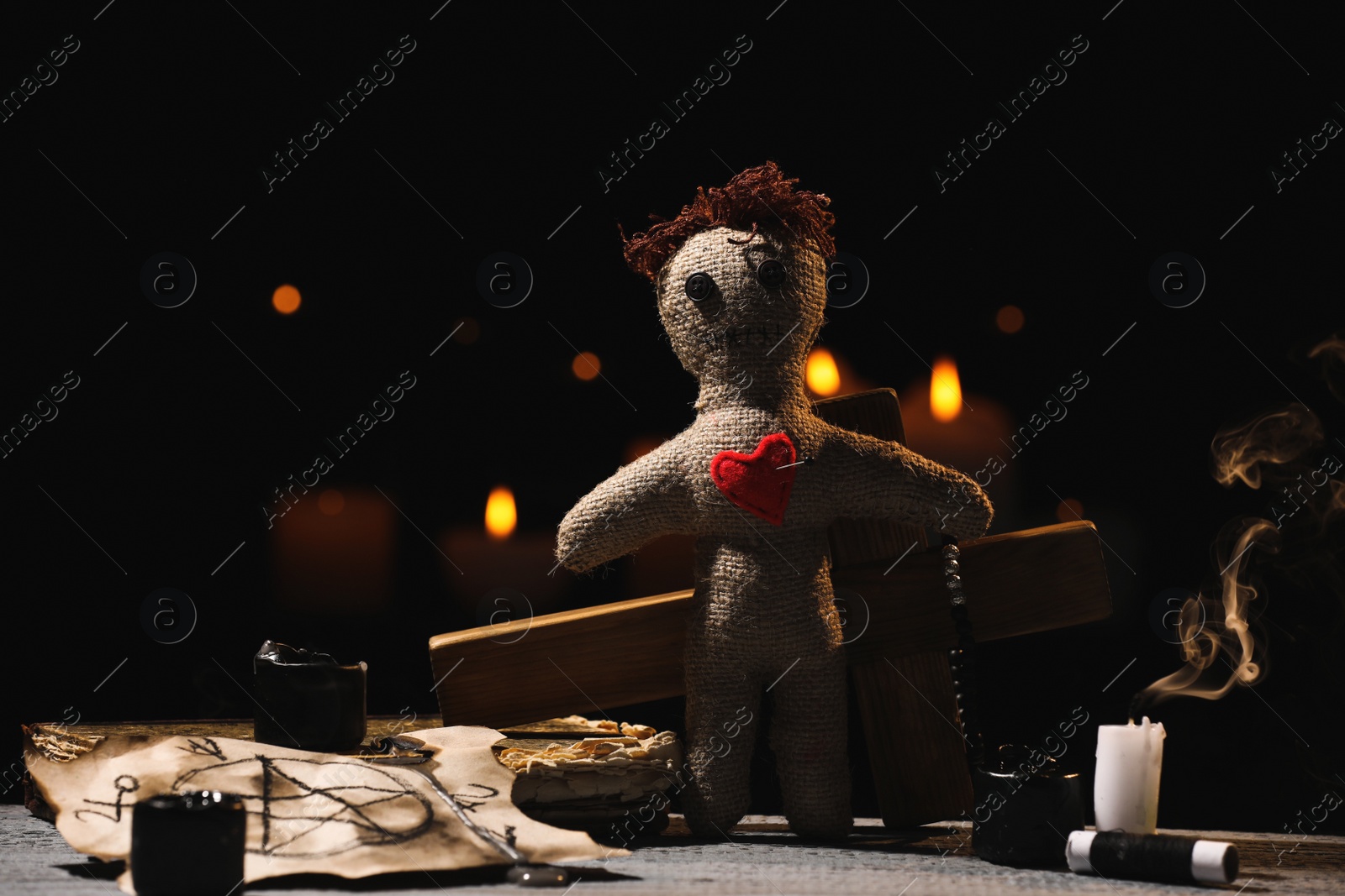 Photo of Voodoo doll with pin in heart and ceremonial items on wooden table against blurred background