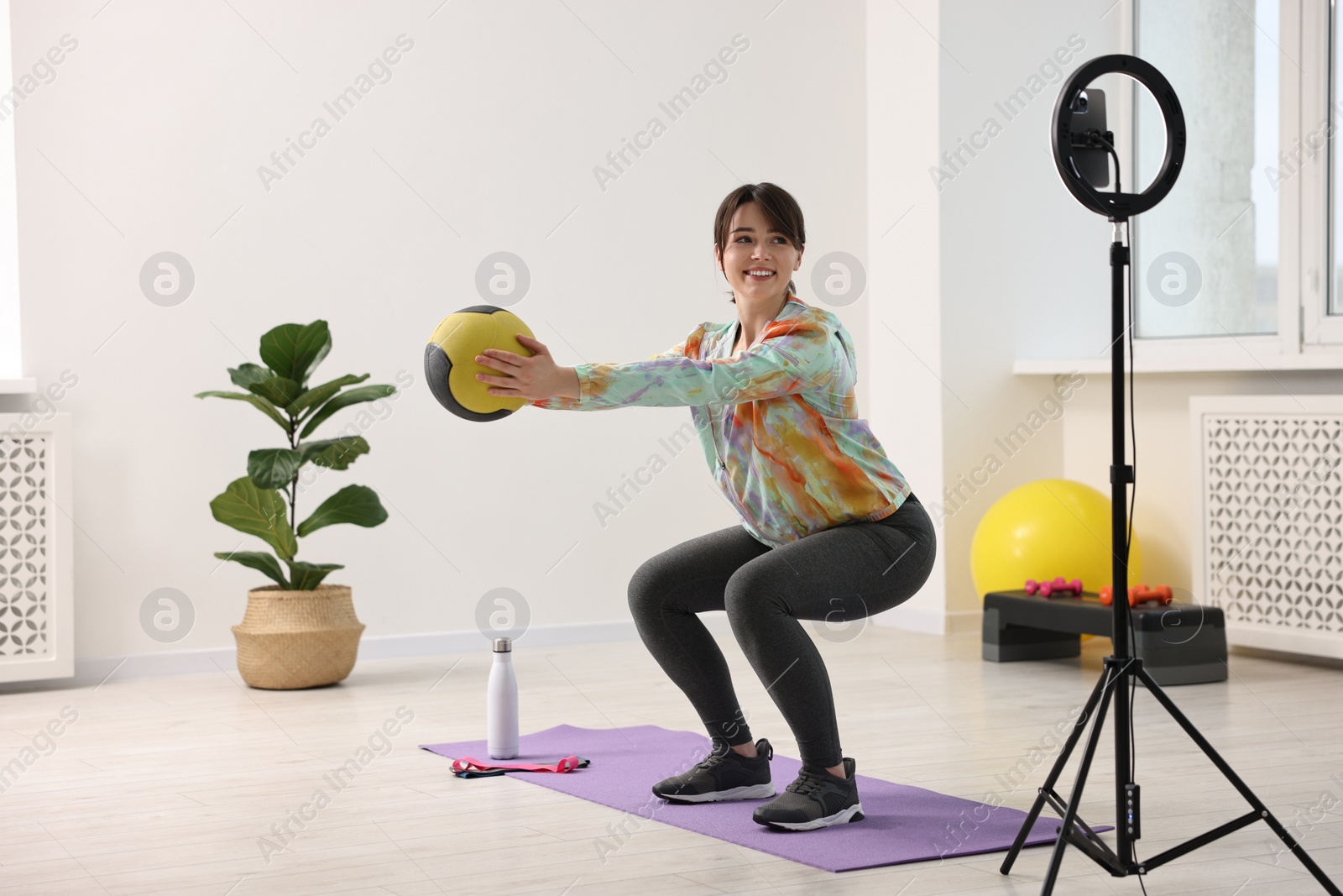 Photo of Happy sports blogger training with medicine ball while streaming online fitness lesson with smartphone at home