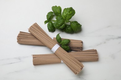 Photo of Uncooked buckwheat noodles (soba) and basil on white marble table, flat lay