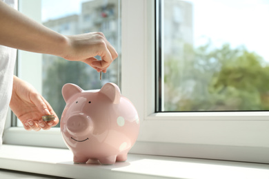 Photo of Woman putting money into piggy bank at window sill indoors, closeup. Space for text