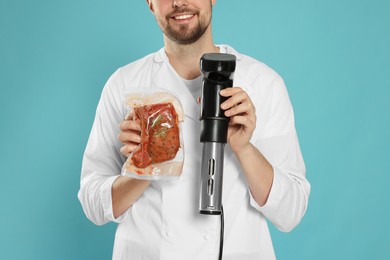 Smiling chef holding sous vide cooker and meat in vacuum pack on light blue background, closeup