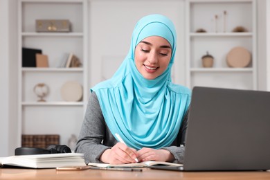 Muslim woman writing notes near laptop at wooden table in room