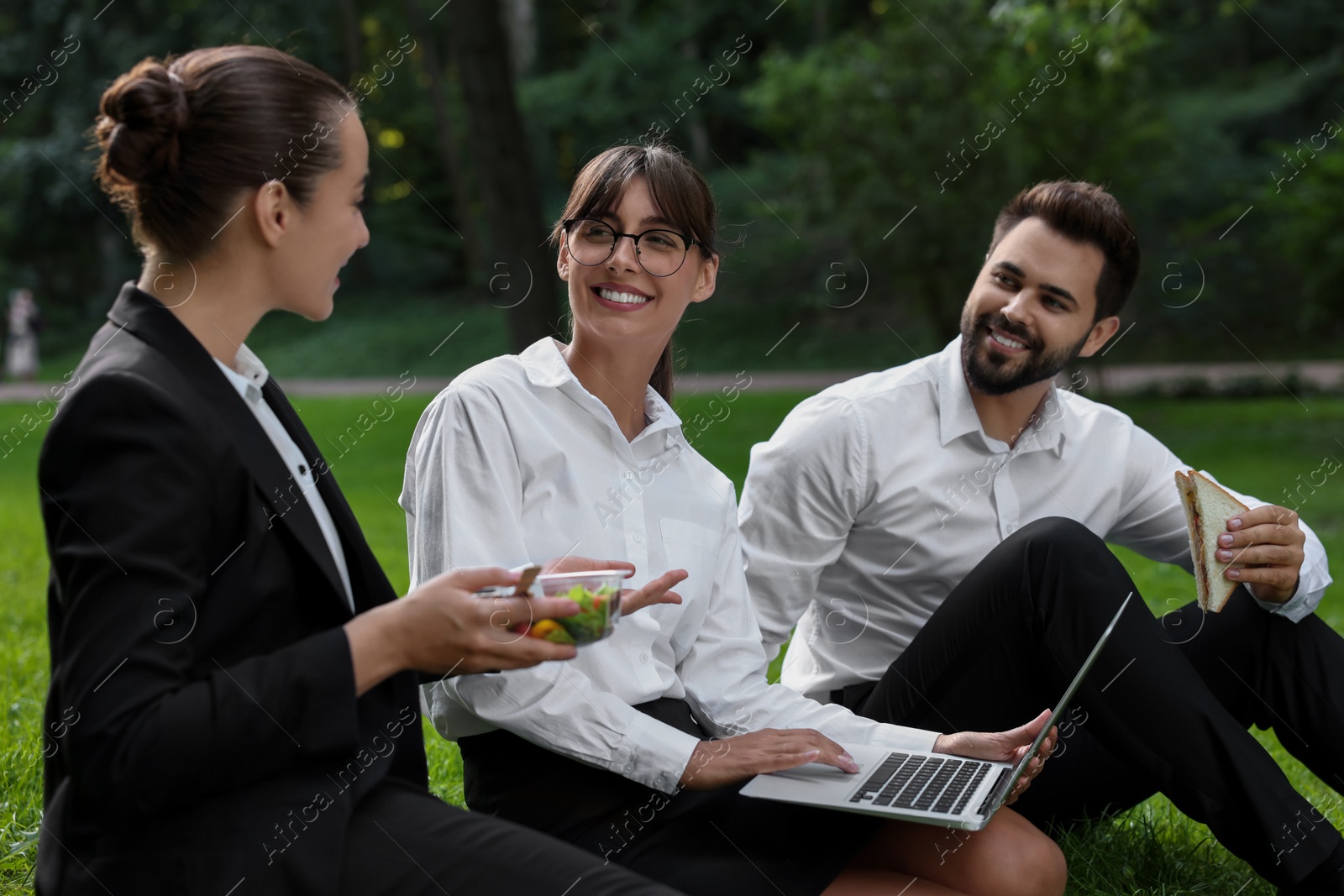 Photo of Happy colleagues with laptop having business lunch on green grass in park