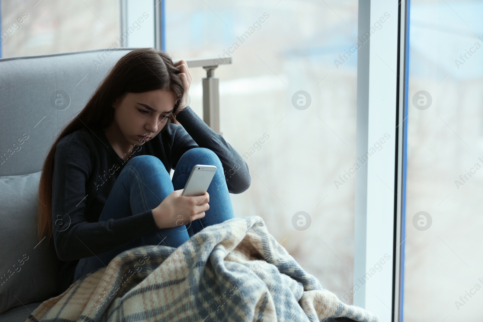 Photo of Upset teenage girl with smartphone sitting at window indoors. Space for text