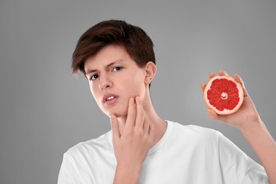 Photo of Teenage boy with acne problem holding grapefruit on grey background. Skin allergy