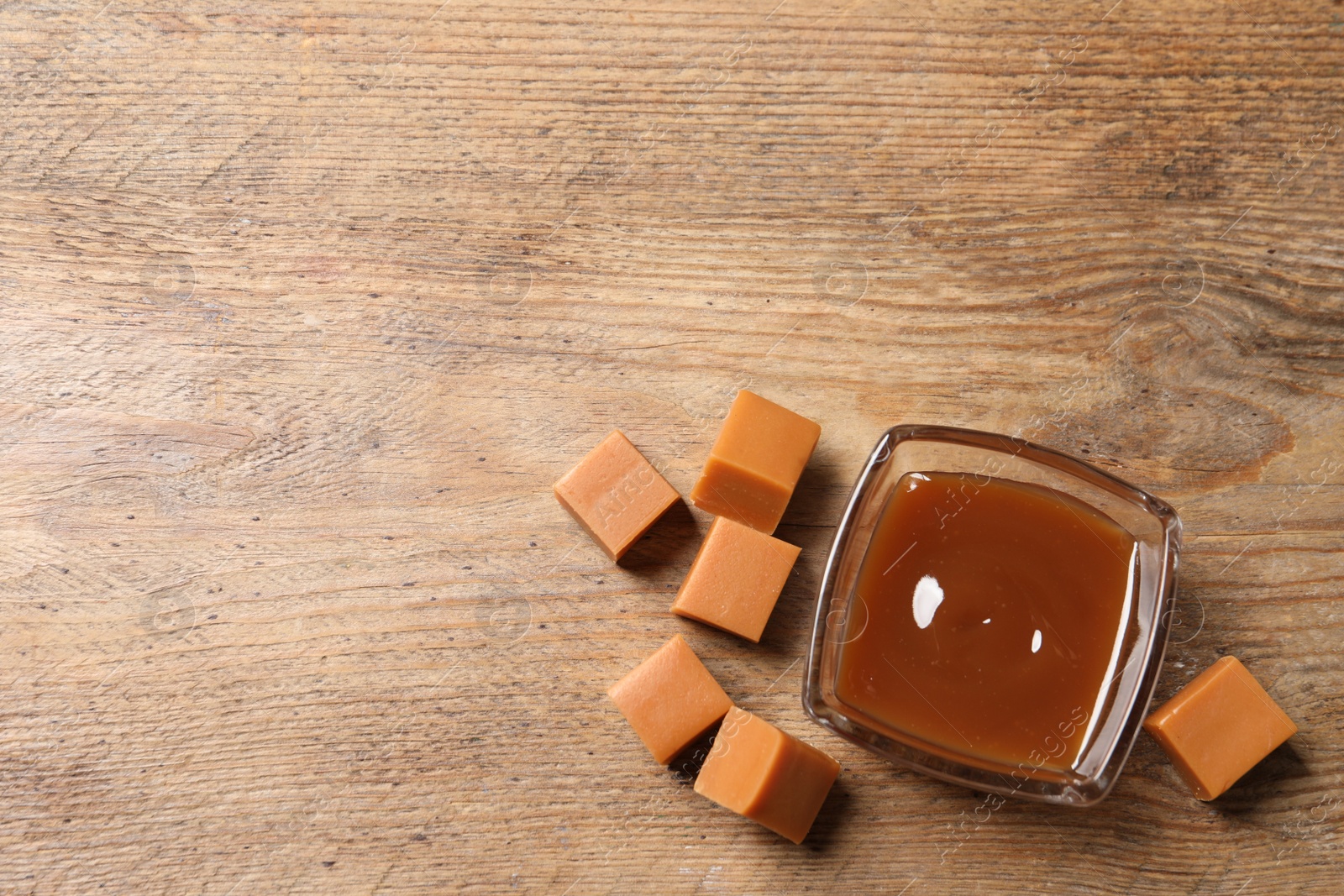 Photo of Tasty salted caramel in glass bowl and candies on wooden table, flat lay. Space for text