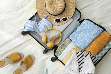 Open suitcase full of clothes, slippers and summer accessories on bed, flat lay