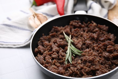 Photo of Fried ground meat in frying pan and rosemary on white tiled table, closeup