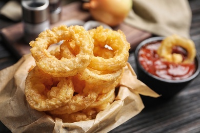Photo of Dishware with homemade crunchy fried onion rings and tomato sauce on wooden table, closeup