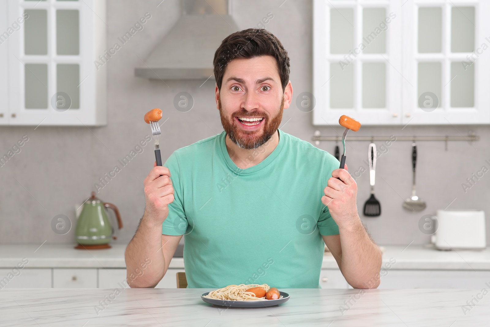 Photo of Man holding forks with sausages and pasta at table in kitchen