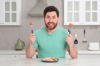 Photo of Man holding forks with sausages and pasta at table in kitchen
