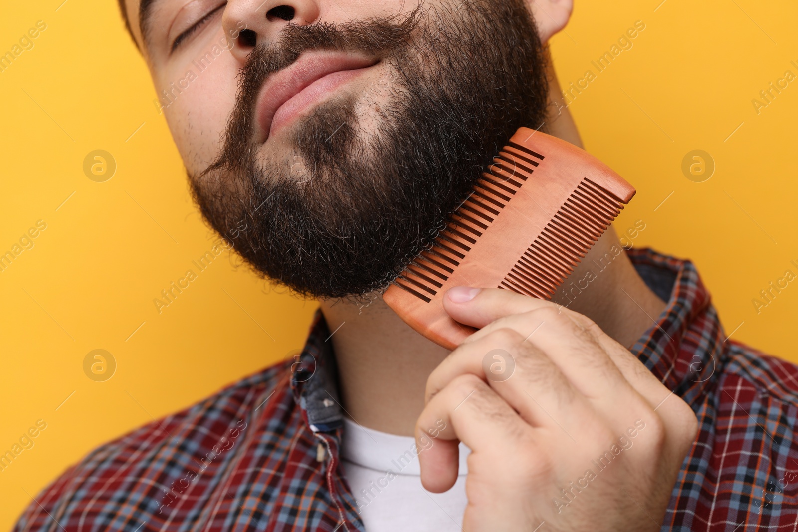 Photo of Handsome young man combing beard on yellow background, closeup