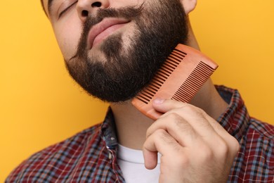 Handsome young man combing beard on yellow background, closeup