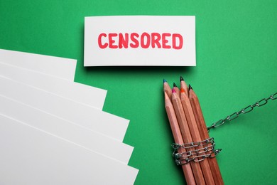 Photo of Card with word Censored, paper sheets, colorful pencils and chain on green background, flat lay