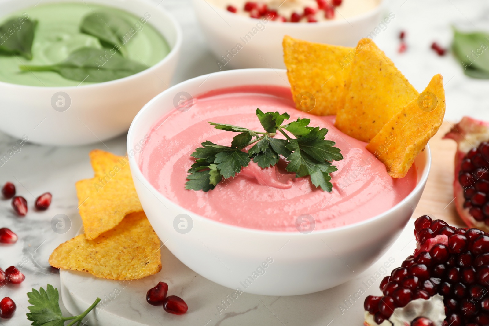 Photo of Bowl of delicious pink hummus with tortilla chips and parsley on white marble table