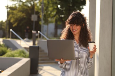 Photo of Happy young woman with cup of coffee using modern laptop outdoors. Space for text