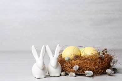 Cute ceramic bunnies near decorative nest with Easter eggs on table against light background