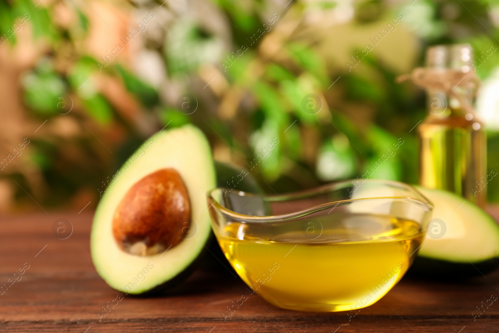 Photo of Glass bowl of cooking oil and fresh avocados on wooden table against blurred green background