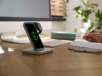 Photo of Woman at desk, smartphone and watch on wireless charger, closeup. Modern workplace