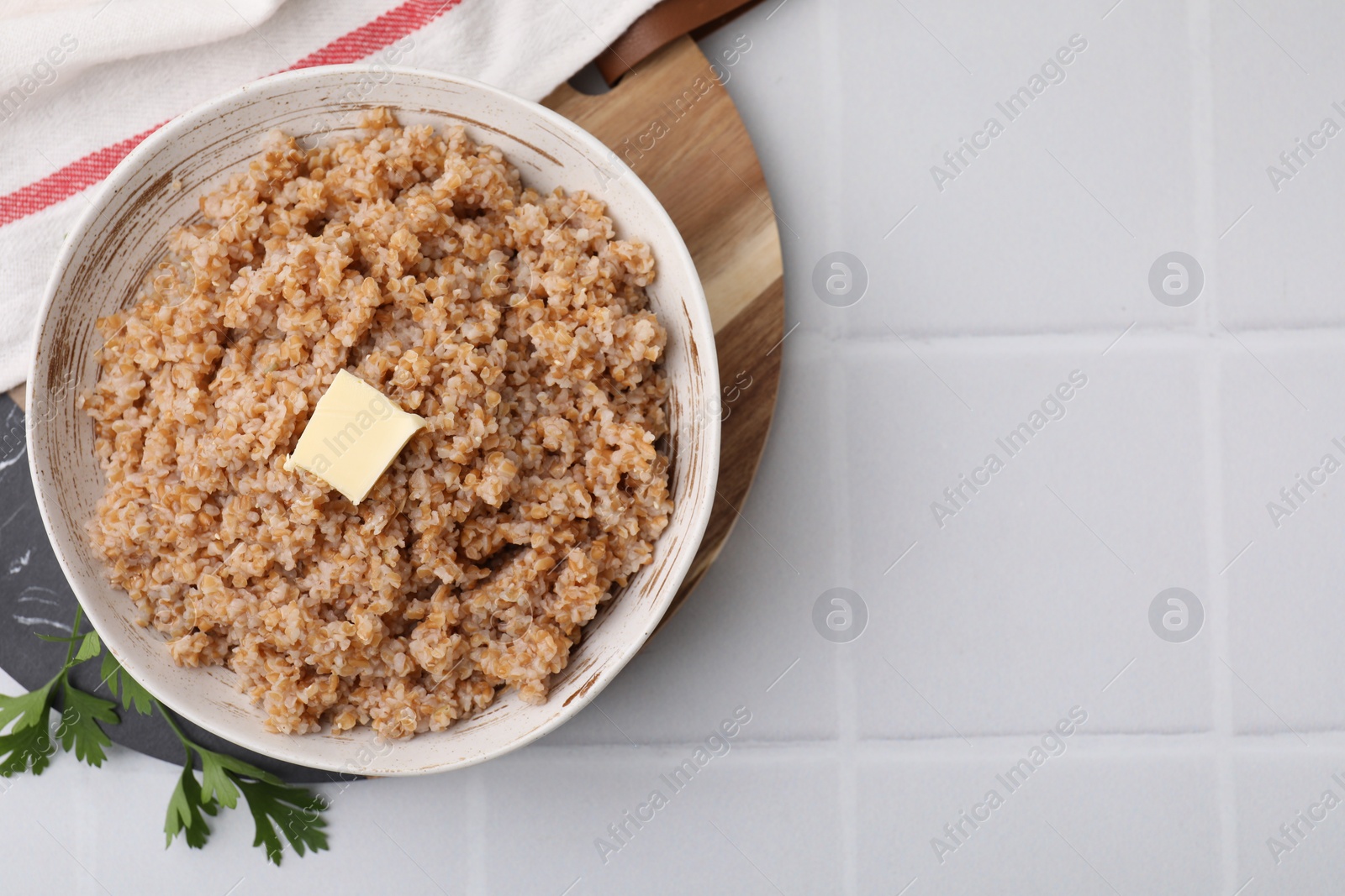 Photo of Tasty wheat porridge with butter and parsley in bowl on white tiled table, top view. Space for text