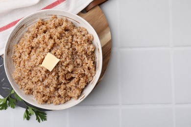 Tasty wheat porridge with butter and parsley in bowl on white tiled table, top view. Space for text