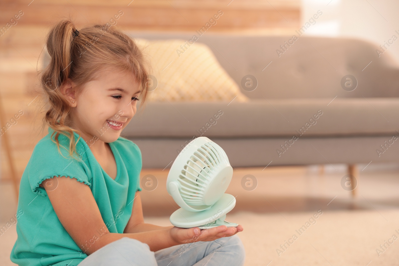 Photo of Little girl enjoying air flow from portable fan in living room. Summer heat