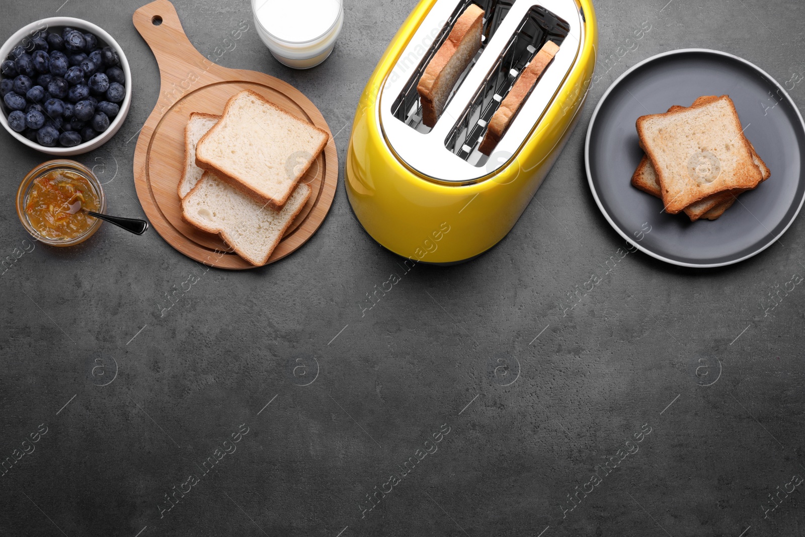 Photo of Yellow toaster with roasted bread, glass of milk, blueberries and jam on grey table, flat lay. Space for text