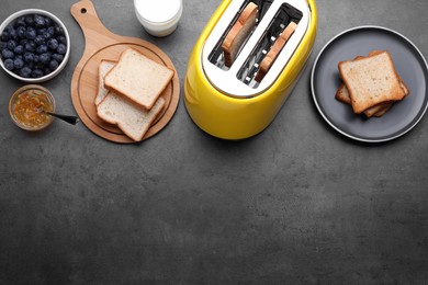 Photo of Yellow toaster with roasted bread, glassmilk, blueberries and jam on grey table, flat lay. Space for text