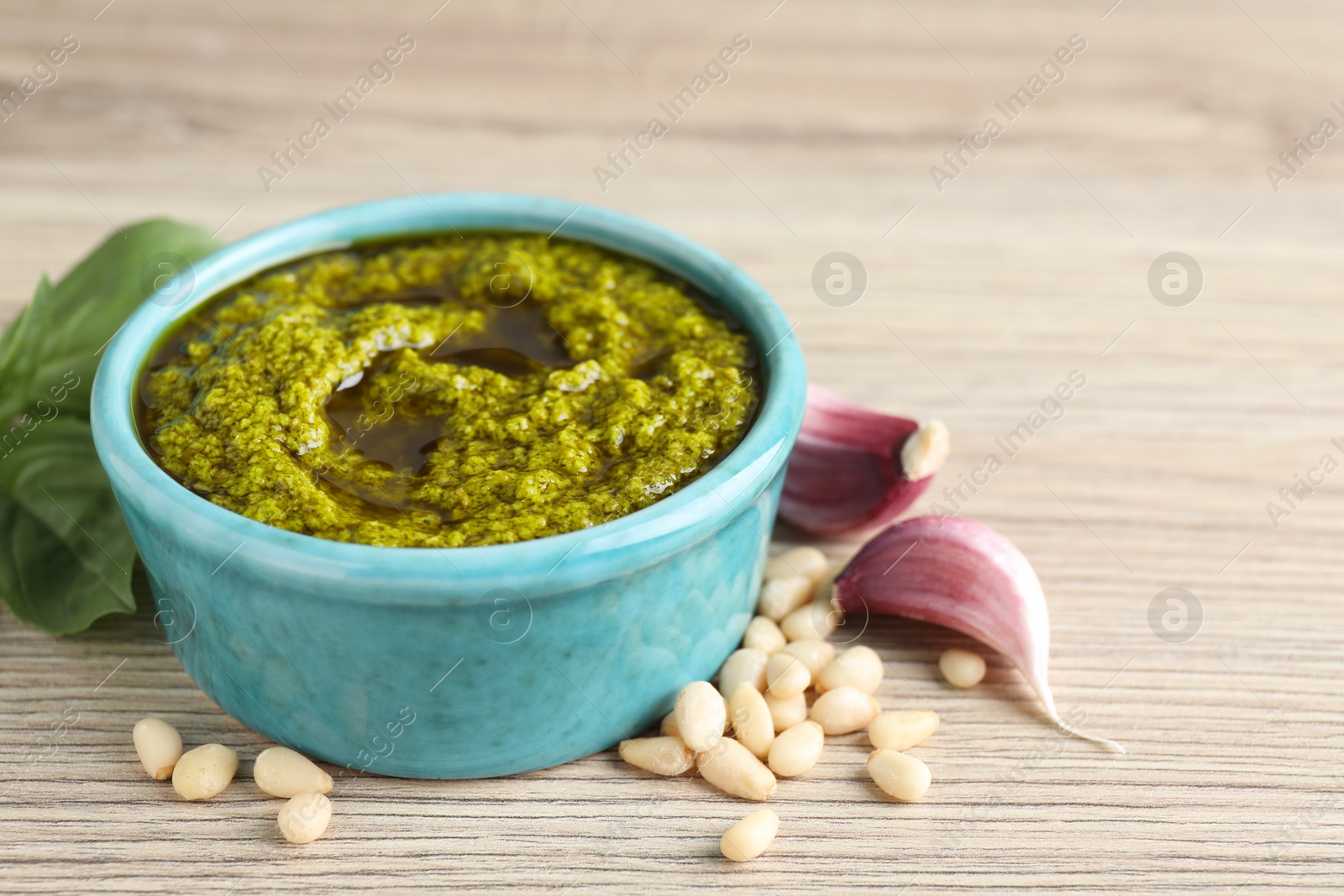 Photo of Tasty pesto sauce in bowl, pine nuts, garlic and basil on wooden table, closeup